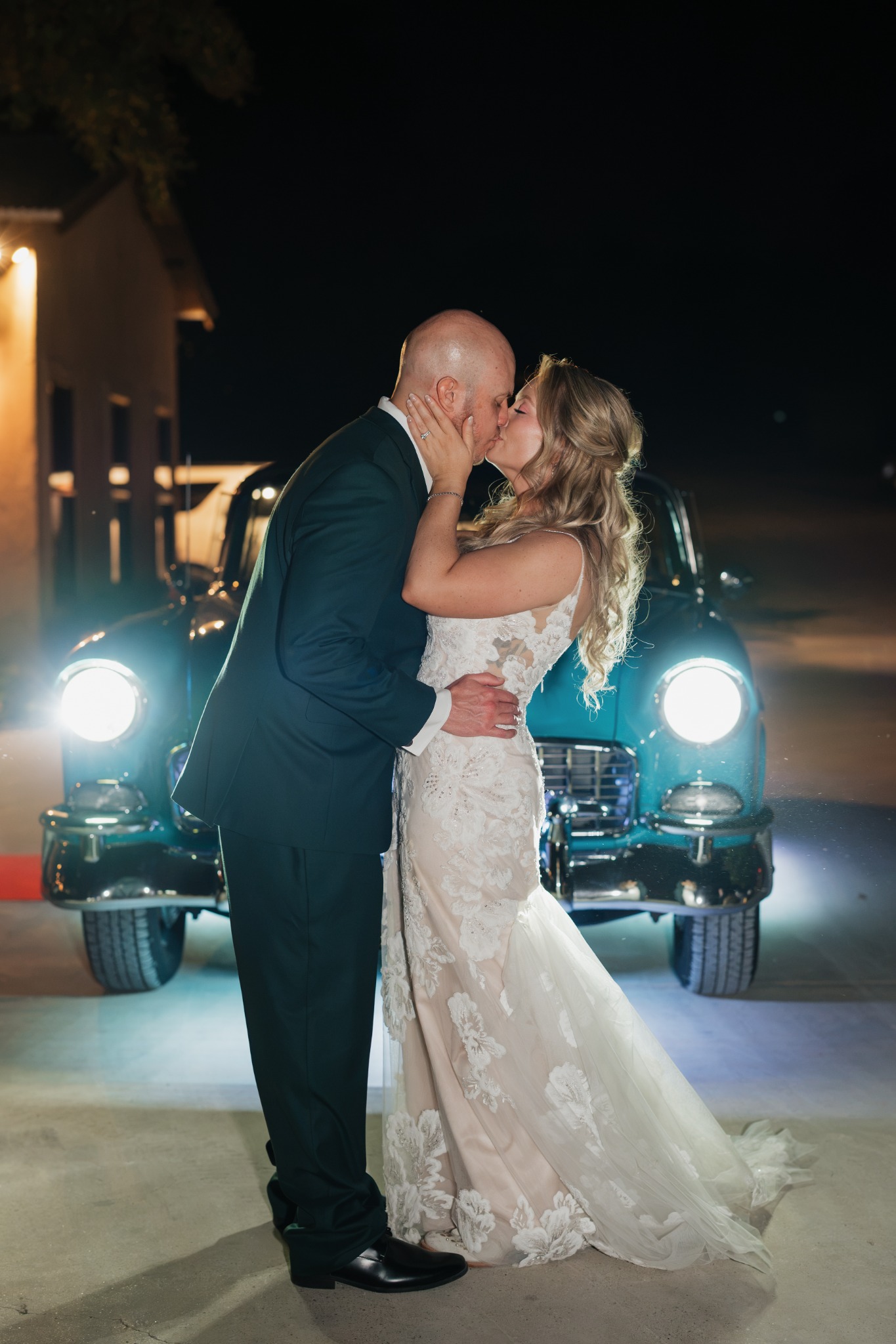 Newlyweds sharing a kiss in front of Ole' Belle with her headlights glowing at night